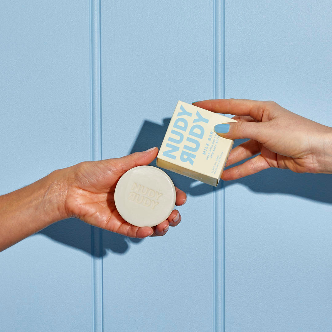 Two hands holding a bar of soap and its packaging against a light blue background